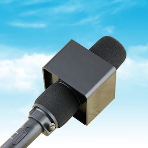Peut inclure: Un microphone noir avec un support carré noir et deux protections en mousse noires. Le microphone est incliné vers le spectateur sur fond de ciel bleu.