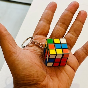 Könnte beinhalten: Ein Miniatur-Rubik's Cube-Schlüsselanhänger mit einem silberfarbenen Ring und einer Kette. Der Würfel hat ein klassisches Farbschema aus Rot, Blau, Grün, Gelb, Orange und Weiß. Der Würfel wird in einer offenen Hand gehalten.