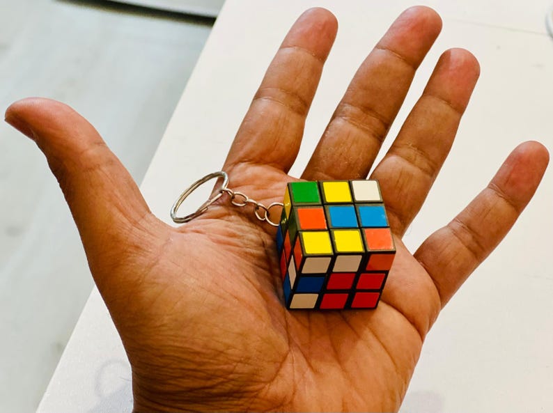 Puede incluir: Un llavero de cubo de Rubik en miniatura con un anillo y una cadena de color plateado. El cubo presenta una combinaci&oacute;n de colores cl&aacute;sica: rojo, azul, verde, amarillo, naranja y blanco. El cubo se sostiene en una mano abierta.