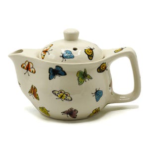 Op de afbeelding: Een witte theepot van keramiek met een deksel, versierd met kleurrijke vlindermotieven. De theepot heeft een tuit en een handvat.