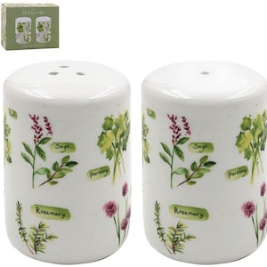 May include: White ceramic salt and pepper shakers decorated with herb illustrations. The shakers display images of sage, parsley, and rosemary, with the herb names printed below. A matching box with the words "Herb Garden" is also visible.