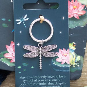 Può includere: Un portachiavi a forma di libellula argentata con un anello argentato. Il portachiavi è su una carta con sfondo blu e una citazione che recita: "Che questo portachiavi a forma di libellula sia un simbolo della tua resilienza, un promemoria costante che nonostante i cambiamenti della vita, la tua forza e la tua bellezza brillano attraverso."