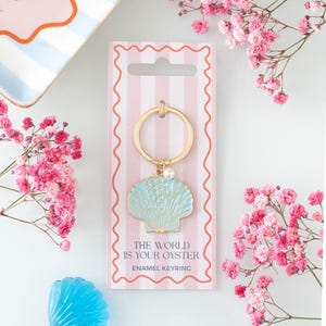 May include: A gold-coloured keyring featuring a light blue seashell enamel charm and a small white pearl. The keyring is attached to a pink and white striped card with the text "THE WORLD IS YOUR OYSTER" and "ENAMEL KEYRING". Pink flowers surround the keyring.