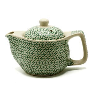 May include: A cream-coloured ceramic teapot with a green floral pattern. The teapot has a matching lid with a small knob and a spout. The handle is rectangular and attached to the side of the pot.