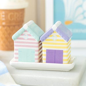 May include: A pair of ceramic beach hut salt and pepper shakers. One is pink and white striped with a mint green door and roof. The other is yellow and white striped with a purple door and roof. They are on a white rectangular tray.
