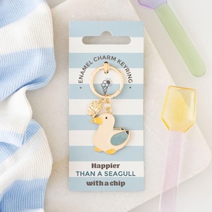 May include: Gold-coloured enamel charm keyring with a duck charm. The duck is cream with blue and yellow details. The keyring is attached to a blue and white striped card with the text "Happier THAN A SEAGULL with a chip."