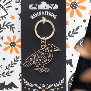 May include: A black metal raven keyring with gold-coloured outlines and decorative floral and star patterns on its body. The raven charm is attached to a gold-coloured metal split ring and is mounted on a black display card labelled RAVEN KEYRING.