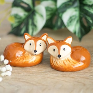 Pode incluir: Um par de saleiros e pimenteiros em cerâmica em forma de raposa. As raposas são laranja com rostos e orelhas creme, e olhos e narizes pretos. Os saleiros estão sobre uma superfície de madeira, com folhagem verde ao fundo.