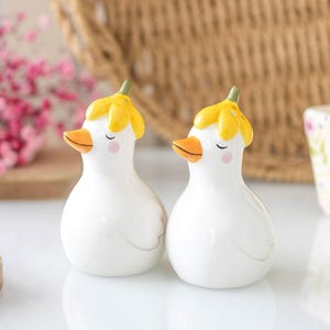 Puede incluir: Dos figuras de patos de cerámica blanca con sombreros amarillos en forma de flor y picos naranjas. Los patos tienen los ojos cerrados y mejillas rosadas. Adecuado para la decoración del hogar.