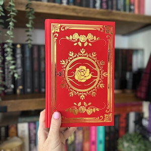 May include: A red book with gold floral embellishments and a central rose design. The cover features intricate gold detailing around the edges and a gold rose in the center. The book is held in front of a bookshelf.