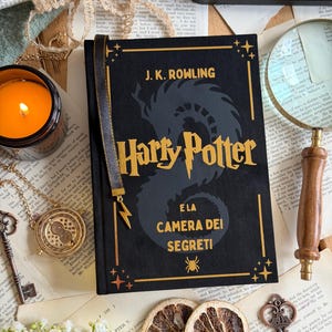 May include: A black hardcover book titled "Harry Potter and the Chamber of Secrets" by J.K. Rowling, with gold lettering and accents. A gold bookmark with a lightning bolt charm is attached. The book is surrounded by vintage-style accessories.