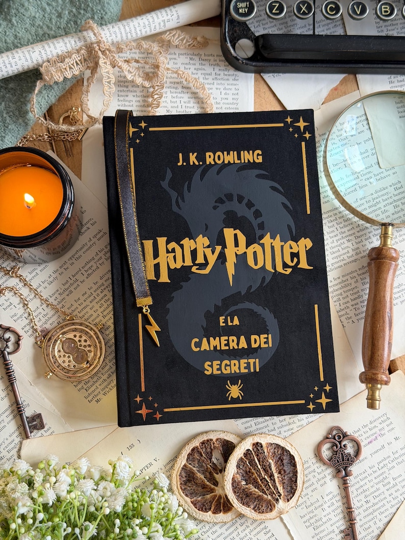 May include: A black hardcover book titled "Harry Potter and the Chamber of Secrets" by J.K. Rowling, with gold lettering and accents. A gold bookmark with a lightning bolt charm is attached. The book is surrounded by vintage-style accessories.