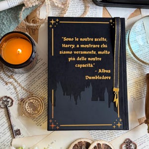 May include: A black journal with gold accents and the quote "Sono le nostre scelte, Harry, a mostrare chi siamo veramente, molto pi&ugrave; delle nostre capacit&agrave;." A gold lightning bolt charm hangs from the bookmark. The journal is surrounded by vintage items.