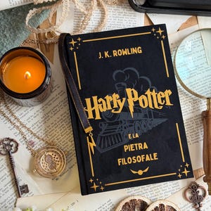 May include: A still-life arrangement featuring a black hardcover book titled "Harry Potter e la Pietra Filosofale" by J.K. Rowling. The book is adorned with gold accents and a train illustration. A lit candle, a necklace, and vintage keys are also present.
