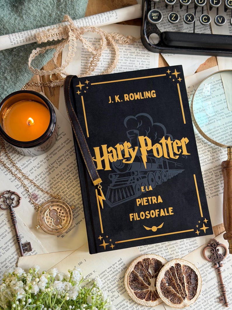 May include: A still-life arrangement featuring a black hardcover book titled "Harry Potter e la Pietra Filosofale" by J.K. Rowling. The book is adorned with gold accents and a train illustration. A lit candle, a necklace, and vintage keys are also present.