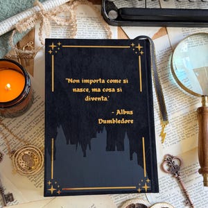 May include: A black book with gold accents and the quote "Non importa come si nasce, ma cosa si diventa" by Albus Dumbledore. The book has a silhouette of a castle and a gold ribbon bookmark.