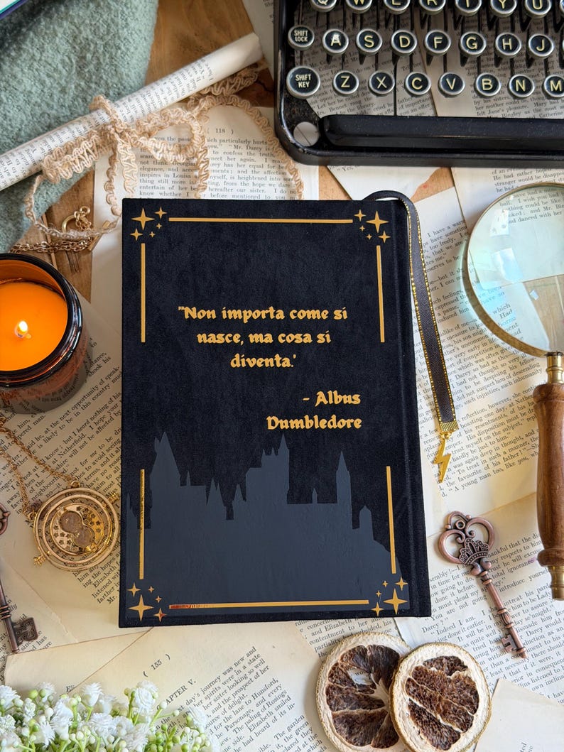 May include: A black book with gold accents and the quote "Non importa come si nasce, ma cosa si diventa" by Albus Dumbledore. The book has a silhouette of a castle and a gold ribbon bookmark.