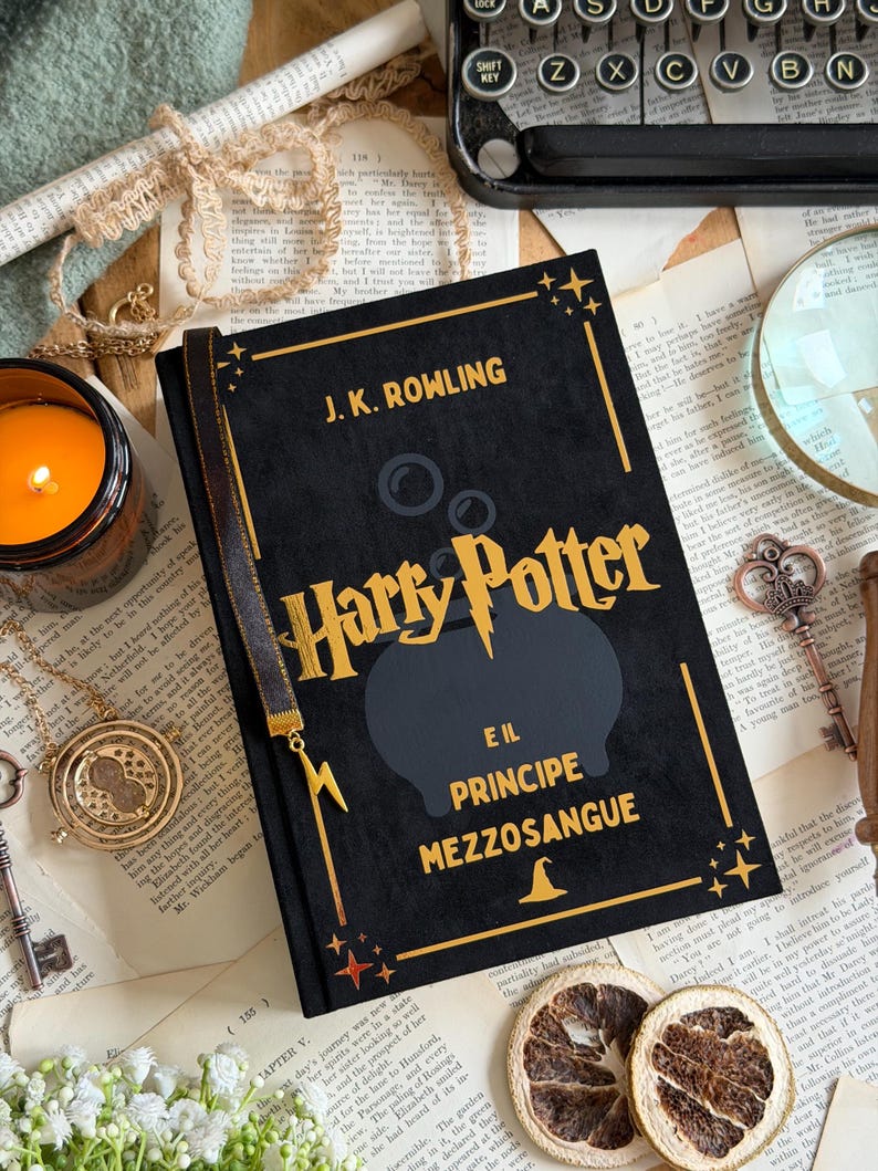 May include: A black hardcover book titled "Harry Potter e il Principe Mezzosangue" by J.K. Rowling. The book has gold lettering and a black cauldron graphic. A gold bookmark and a candle are visible.