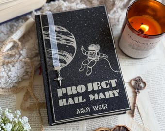 Op de afbeelding: Een zwart boek getiteld "PROJECT HAIL MARY" van Andy Weir, met een ruimtevaartthema-illustratie. Een zilveren lint bladwijzer met een ster charme is ingevoegd. Een brandende kaars en gedroogde sinaasappelschijfjes zijn ook zichtbaar.