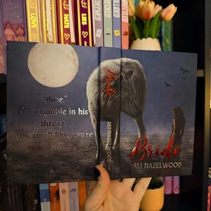 Może przedstawiać: Okładka książki "Bride" autorstwa Ali Hazelwood, przedstawiająca wilka i sylwetkę kobiety pod pełnią księżyca. Tytuł jest w czerwonym skrypcie. Tło to mroczny krajobraz z cytatami z książki.