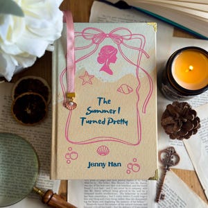 Può includere: Un libro con copertina rigida intitolato "The Summer I Turned Pretty" di Jenny Han. Il libro ha una copertina beige chiaro con illustrazioni e testo rosa. È inserito un segnalibro a nastro rosa con un ciondolo a forma di conchiglia. L'immagine include una candela accesa, una chiave e fette d'arancia essiccate.
