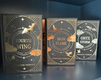Puede incluir: Tres libros de tapa dura con cubiertas oscuras y detalles dorados. Los títulos son "Fourth Wing", "Iron Flame" y "Onyx Storm" de Rebecca Yarros. Cada libro presenta un diseño circular con dragones, estrellas y nubes.