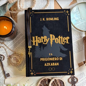 May include: A black hardcover book titled "Harry Potter and the Prisoner of Azkaban" with gold lettering and a silhouette of a wolf. A gold lightning bolt bookmark is attached. The book is surrounded by vintage-style decor.