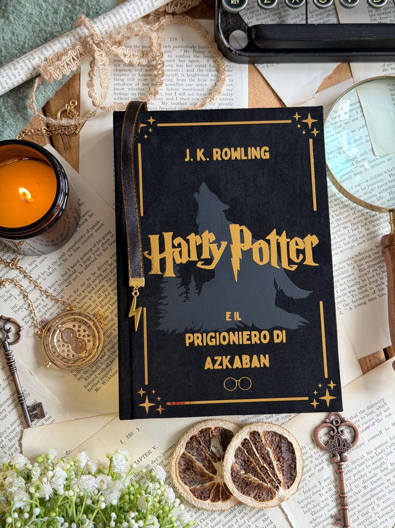 May include: A black hardcover book titled "Harry Potter and the Prisoner of Azkaban" with gold lettering and a silhouette of a wolf. A gold lightning bolt bookmark is attached. The book is surrounded by vintage-style decor.