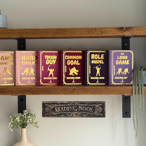 Op de afbeelding: Zes hardcover boeken met gouden letters en illustraties worden tentoongesteld op een houten plank. De titels zijn "Game Changer", "Heated Rivalry", "Tough Guy", "Common Goal", "Role Model" en "The Long Game". Een "Reading Nook" bordje staat eronder.