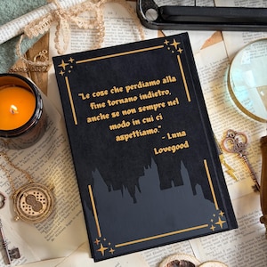 May include: A black journal with gold accents and the quote "Le cose che perdiamo alla fine tornano indietro, anche se non sempre nel modo in cui ci aspettiamo." by Luna Lovegood. The journal is surrounded by vintage items like a typewriter and a candle.