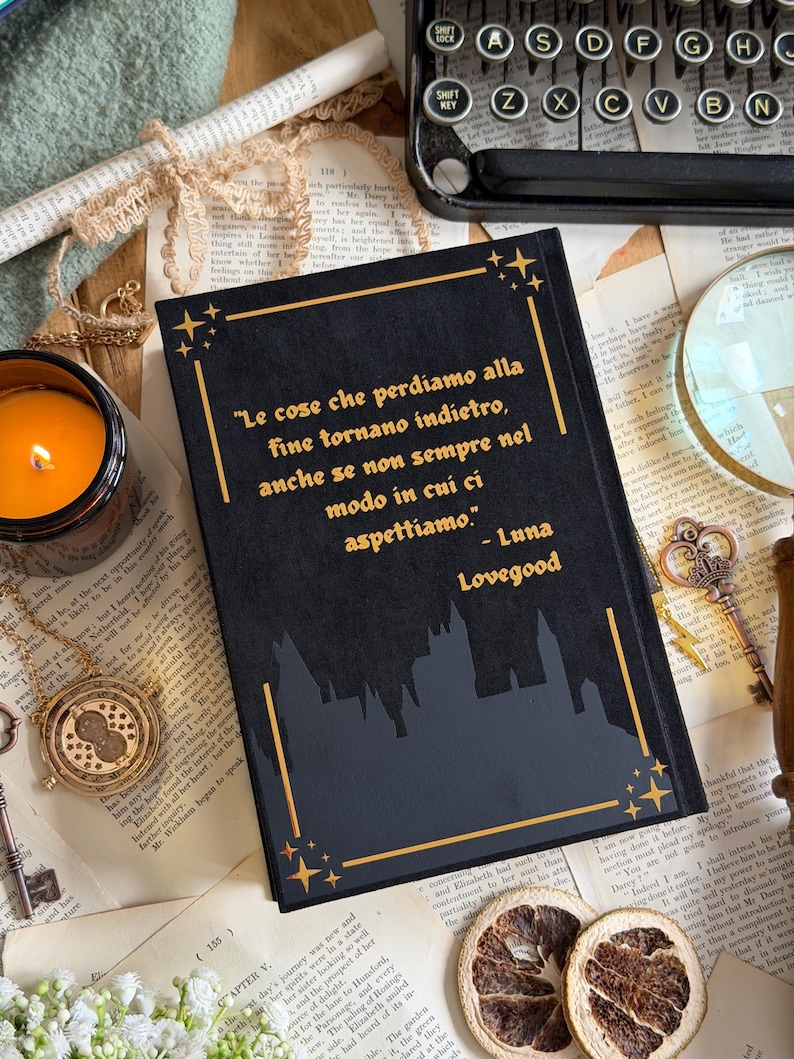 May include: A black journal with gold accents and the quote "Le cose che perdiamo alla fine tornano indietro, anche se non sempre nel modo in cui ci aspettiamo." by Luna Lovegood. The journal is surrounded by vintage items like a typewriter and a candle.