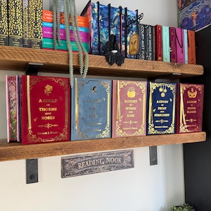 Op de afbeelding: Twee houten planken met een boekenverzameling. De onderste plank bevat vijf boeken met goudkleurige titels, waaronder A Court of Thorns and Roses, A Court of Mist and Fury, A Court of Wings and Ruin, A Court of Frost and Starlight en A Court of Silver Flames. Een bordje eronder vermeldt Reading Nook.