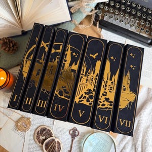May include: A collection of seven black book boxes with gold foil designs and Roman numerals I through VII. The boxes feature castle and bird silhouettes. Other items include a vintage typewriter, magnifying glass, and dried orange slices.