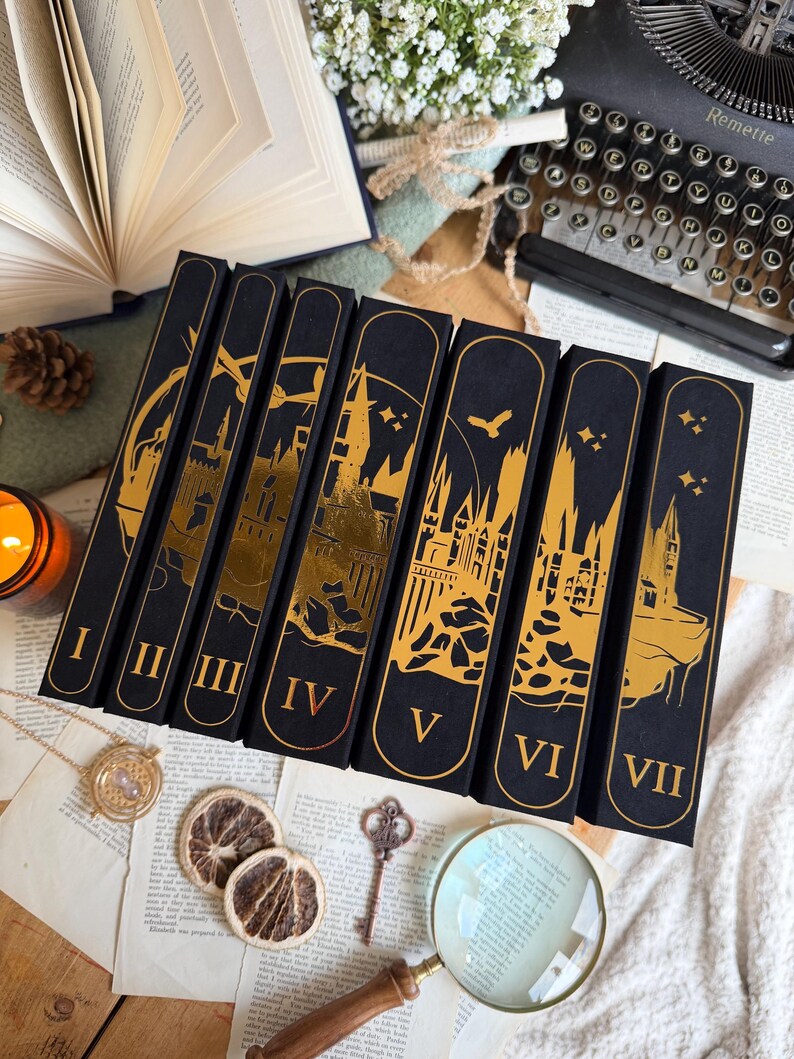 May include: A collection of seven black book boxes with gold foil designs and Roman numerals I through VII. The boxes feature castle and bird silhouettes. Other items include a vintage typewriter, magnifying glass, and dried orange slices.