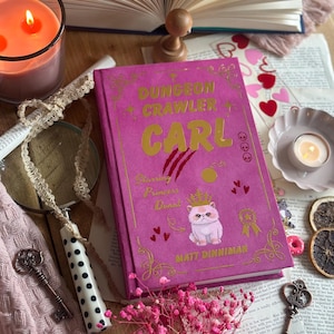 Op de afbeelding: Een roze hardcover boek met de titel "DUNGEON CRAWLER CARL" in gouden letters en een kattenillustratie. De afbeelding bevat een brandende kaars, sleutels en gedroogde bloemen. De naam van de auteur, Matt Dinniman, is zichtbaar.