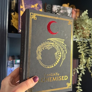 May include: A dark gray book with gold foil accents and the title "ALCHEMISED". The cover features a gold dragon design and a red crescent moon. The book is held in front of a dark blue bookshelf.