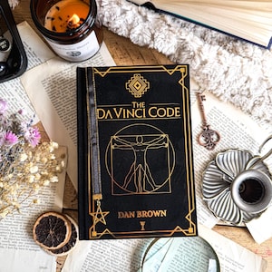 Pode incluir: Um livro de capa dura preto intitulado The Da Vinci Code de Dan Brown com letras douradas e uma ilustração do Homem Vitruviano na capa. O livro possui um marcador de fita preta com um pingente de estrela dourada na extremidade.