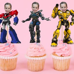 Pode incluir: Três toppers para cupcake com personagens de Transformers de desenhos animados com rostos humanos. Os personagens são Optimus Prime, Bumblebee e Ironhide. Os cupcakes são cobertos com glacê rosa com granulado azul e branco.