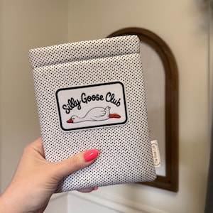 May include: A white and black polka dot book sleeve with a patch that reads "Silly Goose Club" featuring a cartoon goose. The sleeve is held by a person with pink nail polish. A small tag reads "The Book Loft".