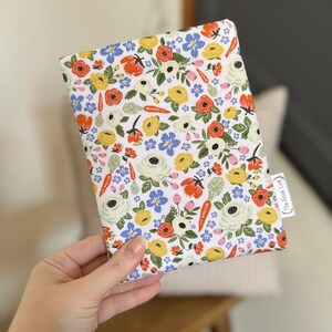 K&ouml;nnte beinhalten: Eine wei&szlig;e Stofftasche mit einem Blumenmuster mit roten, gelben und blauen Blumen, gr&uuml;nen Bl&auml;ttern und orangefarbenen Karotten. Die Tasche hat ein kleines Etikett mit der Aufschrift "The Book Lady".