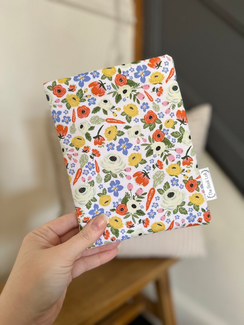 K&ouml;nnte beinhalten: Eine wei&szlig;e Stofftasche mit einem Blumenmuster mit roten, gelben und blauen Blumen, gr&uuml;nen Bl&auml;ttern und orangefarbenen Karotten. Die Tasche hat ein kleines Etikett mit der Aufschrift "The Book Lady".