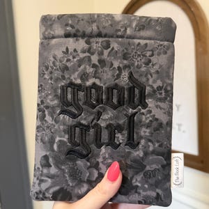 May include: A gray floral book sleeve with the embroidered text "GOOD GIRL" in black gothic lettering. The sleeve is held by a hand with a pink nail polish. A small white tag with text is visible.