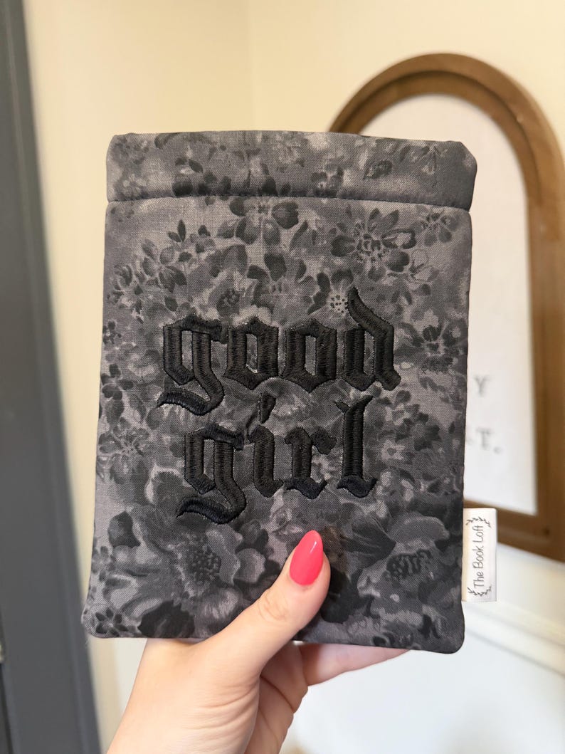 May include: A gray floral book sleeve with the embroidered text "GOOD GIRL" in black gothic lettering. The sleeve is held by a hand with a pink nail polish. A small white tag with text is visible.