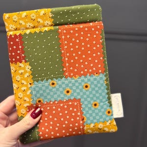 May include: A patchwork fabric case with a variety of patterns and colors. The case features squares of orange, green, yellow, and blue fabric. Some squares have floral patterns, while others have polka dots or sunflowers. A small white tag is attached to the side.