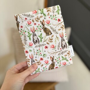 K&ouml;nnte beinhalten: Eine wei&szlig;e Stofftasche mit einem sich wiederholenden Muster aus braunen Kaninchen, gr&uuml;nen Bl&auml;ttern und rosa Bl&uuml;ten. Die Tasche hat ein kleines wei&szlig;es Etikett mit dem Text "The Bookish Owl" darauf.