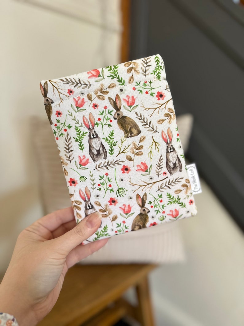 K&ouml;nnte beinhalten: Eine wei&szlig;e Stofftasche mit einem sich wiederholenden Muster aus braunen Kaninchen, gr&uuml;nen Bl&auml;ttern und rosa Bl&uuml;ten. Die Tasche hat ein kleines wei&szlig;es Etikett mit dem Text "The Bookish Owl" darauf.