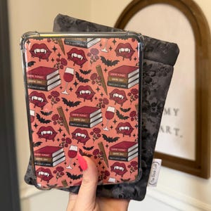 May include: A clear protective case with a pink background featuring a pattern of vampire fangs, bats, books, roses, and wine glasses. A dark gray fabric book sleeve is behind the case.