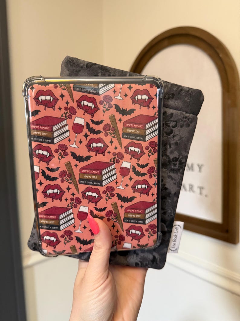 May include: A clear protective case with a pink background featuring a pattern of vampire fangs, bats, books, roses, and wine glasses. A dark gray fabric book sleeve is behind the case.