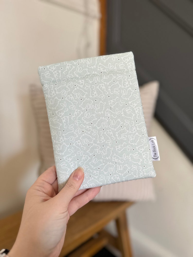 K&ouml;nnte beinhalten: Eine hellgraue Stofftasche mit einem wei&szlig;en Hasenmuster und dem Text "The Book Loft" in der unteren rechten Ecke.