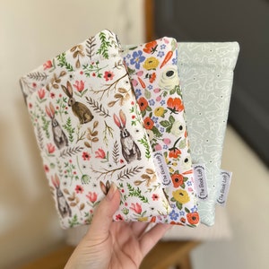 K&ouml;nnte beinhalten: Drei Stoffbeutel mit Blumen- und Hasenmustern. Die Beutel sind wei&szlig; mit verschiedenen farbigen Blumendesigns und haben ein wei&szlig;es Hasenmuster auf der R&uuml;ckseite. Jeder Beutel hat ein wei&szlig;es Etikett mit dem aufgedruckten Text "The Book Loft".